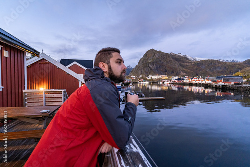 young man with beard drinking from the mug in the bay of Sørvågen typical norwegian village during sunrise on lofoten islands