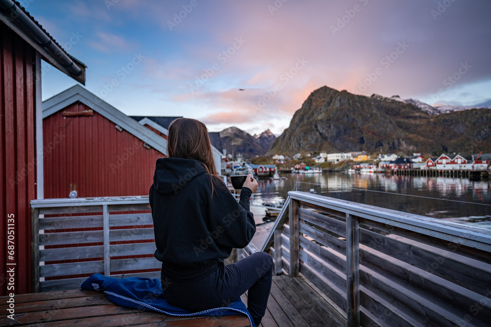 young woman drinking coffee in the morning and taking a look on the bay in Sørvågen fisherman village with red houses on the coast, lofoten islands, norway