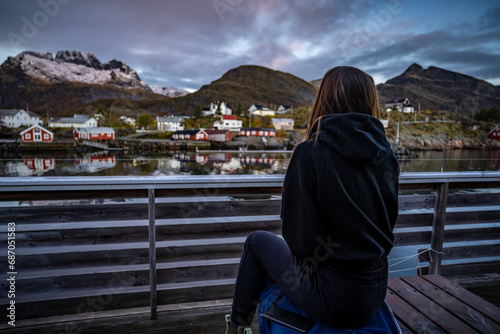 young woman drinking coffee in the morning and taking a look on the bay in Sørvågen fisherman village with red houses on the coast, lofoten islands, norway