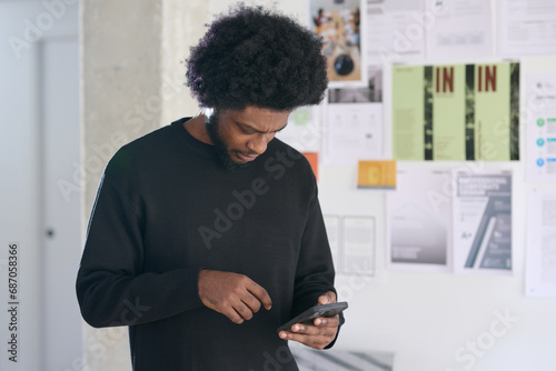 Focused black and afro man sending a text message in the office. Moodboard of varied business projects in the background photo