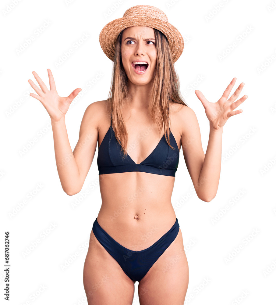 Young beautiful girl wearing bikini and hat crazy and mad shouting and yelling with aggressive expression and arms raised. frustration concept. Young beautiful girl wearing bikini and hat crazy and mad shouting and yelling with aggressive expression and arms raised. frustration concept.