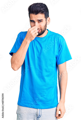 Young handsome man with beard wearing casual t-shirt smelling something stinky and disgusting, intolerable smell, holding breath with fingers on nose. bad smell