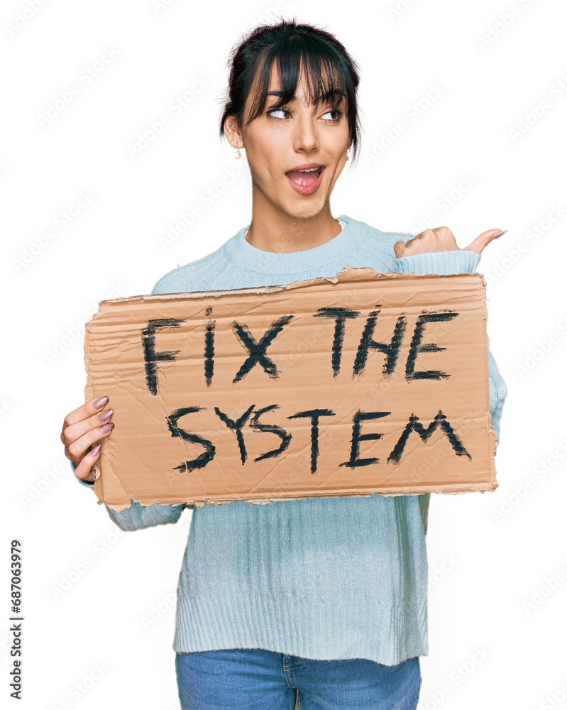 Young hispanic woman holding fix the system banner cardboard pointing thumb up to the side smiling happy with open mouth Young hispanic woman holding fix the system banner cardboard pointing thumb up to the side smiling happy with open mouth