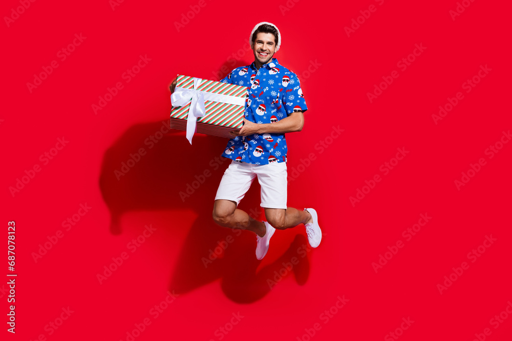 Full body photo of handsome young guy jump hold big present box dressed santa claus print x-mas outfit isolated on red color background Full body photo of handsome young guy jump hold big present box dressed santa claus print x-mas outfit isolated on red color background