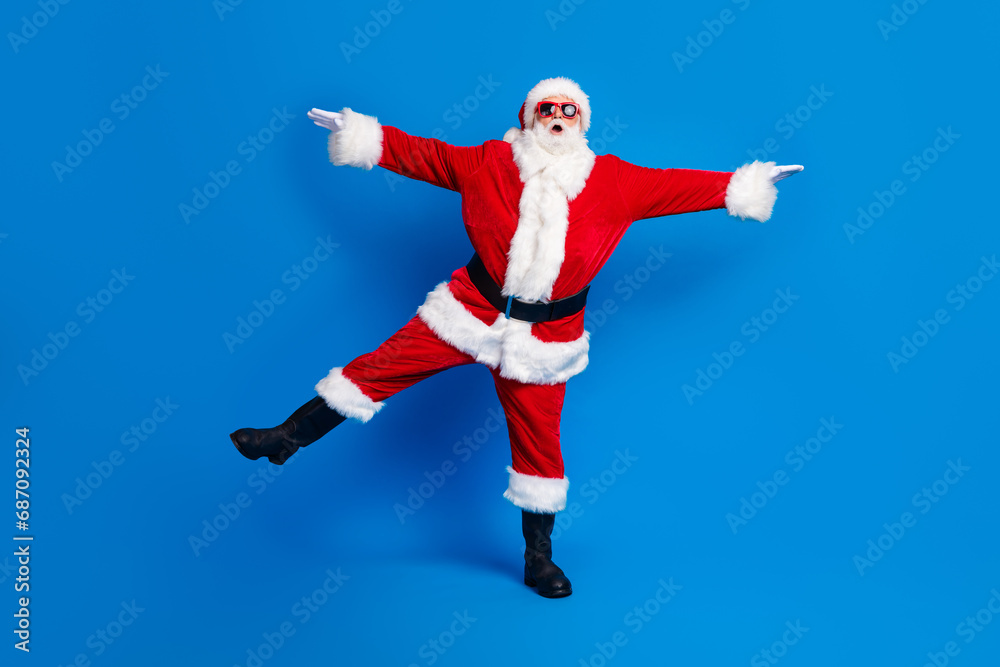 Full size photo of pensioner senior man stretch hands legs have fun wear trendy santa claus costume coat isolated on blue color background Full size photo of pensioner senior man stretch hands legs have fun wear trendy santa claus costume coat isolated on blue color background