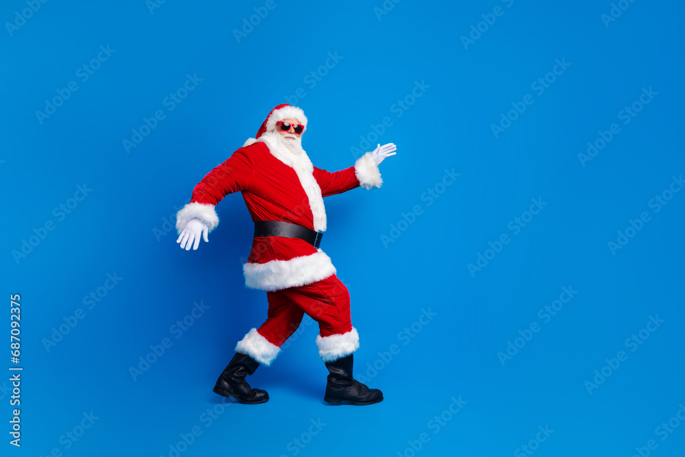 Full length photo of mature pensioner man sneak go deliver gifts wear trendy santa claus costume coat isolated on blue color background Full length photo of mature pensioner man sneak go deliver gifts wear trendy santa claus costume coat isolated on blue color background