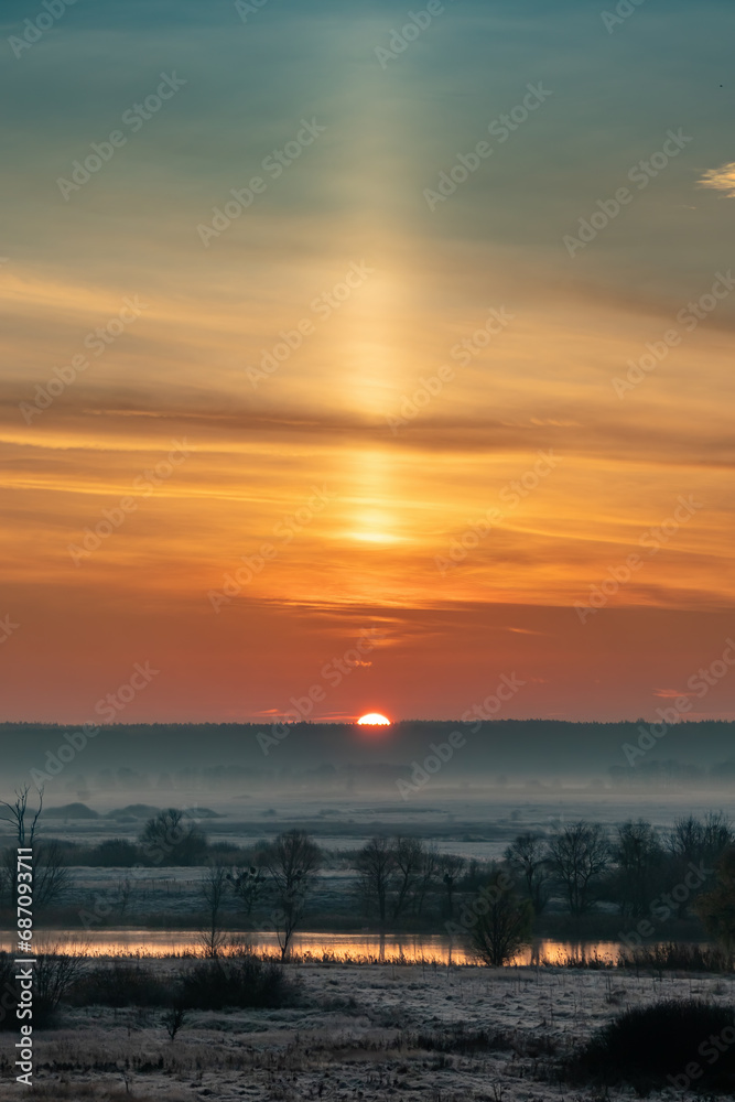 Sunrise over a river valley with trees and meadows on a foggy morning. A light pillar is an atmospheric optical phenomenon, a vertical beam of light from the Sun. Landscape of late autumn or winter