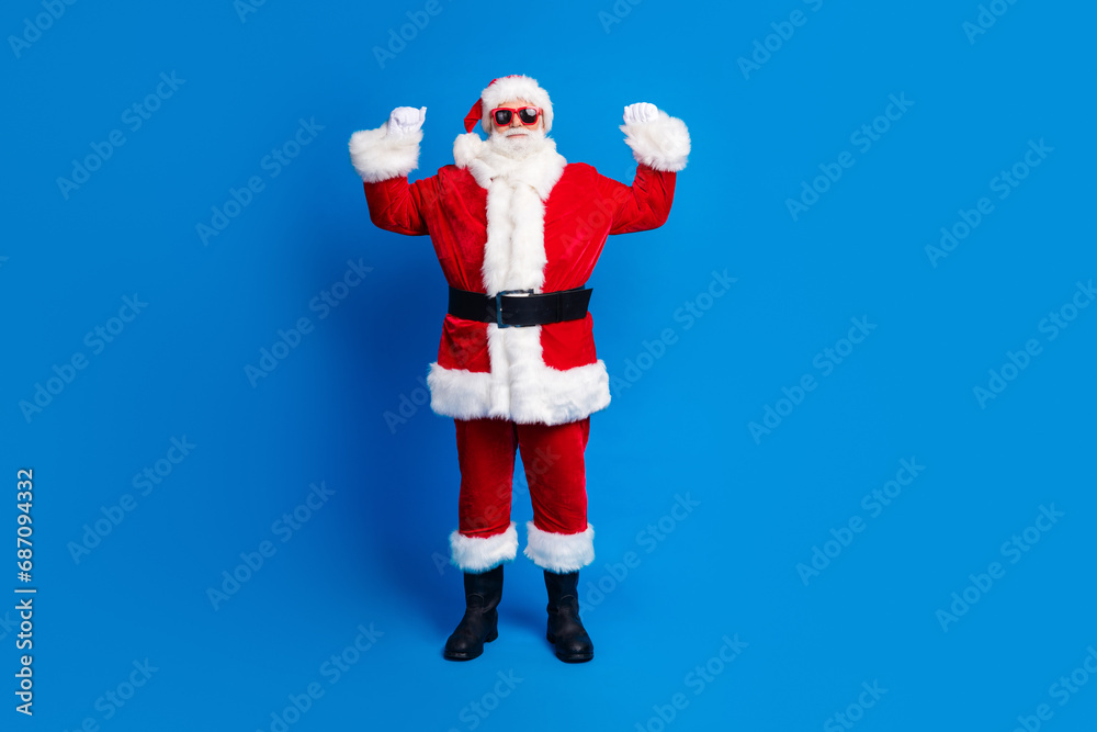 Full size photo of pensioner senior man raise fists victory lottery wear trendy santa claus costume coat isolated on blue color background Full size photo of pensioner senior man raise fists victory lottery wear trendy santa claus costume coat isolated on blue color background