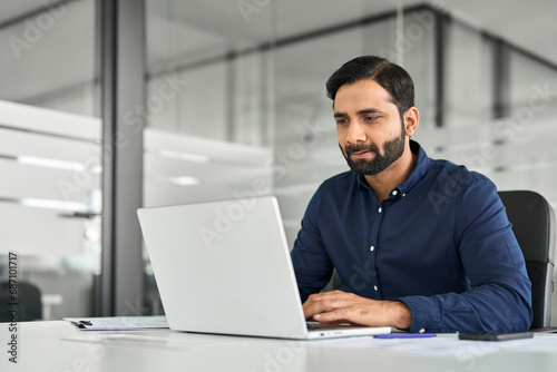 Busy smart Indian business man employee looking at laptop computer, businessman office worker or company manager analyzing project strategy working with ai software sitting at work desk.