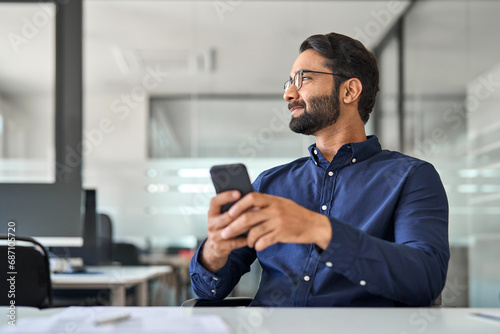 Smiling professional Indian businessman employee looking aside holding smartphone sitting at desk. Happy busy business man executive using cell phone thinking of new mobile solutions working in office