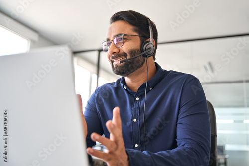 Happy Indian call center agent wearing headset talking to client working in customer support office. Professional contract service telemarketing operator using laptop having conversation. Candid shot photo