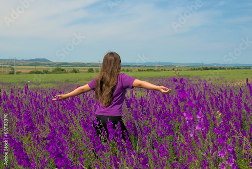 A girl with brown long hair enjoys the scents of spring against the background of a field of purple Delphínium flowers.