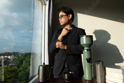asian man wear black suit dark shade glasses stand in fornt of desk books make confidence mood and coffee with city window view and shadow on wall