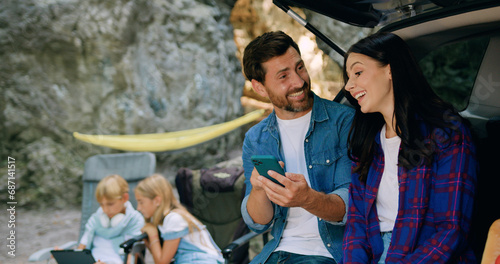 Joyful young adult couple man and woman sitting in car trunk with children on background using tapping on phone while laughing. Mother and father surfing internet on cellphone. Family road trip with