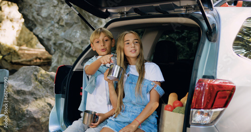 Cheerful brother and sister sitting in the car trunk and drinking tea and speaking during spring hiking trip. Happy family resting outdoor in summer forest. Family road trip with trailer car. © serg