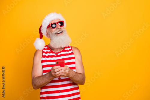 Photo of positive dreamy senior guy christmas swimwear hat glasses typing modern device empty space isolated yellow color background