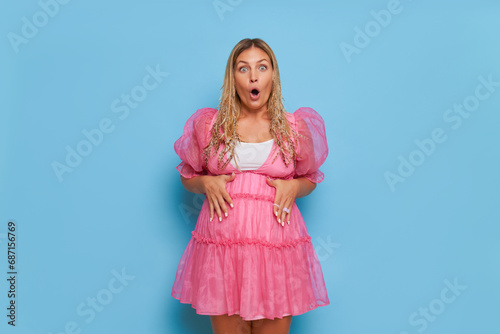 Young pretty expectant mother posing in pink dress on blue background, holding her hands on her stomach with open mouth, good mood concept, copy space