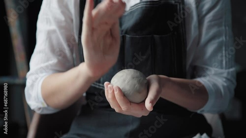 A woman in a black apron holds white clay in hands, makes a ball out of it, and warms it in palms. Medium shot, only the hands in the frame. photo