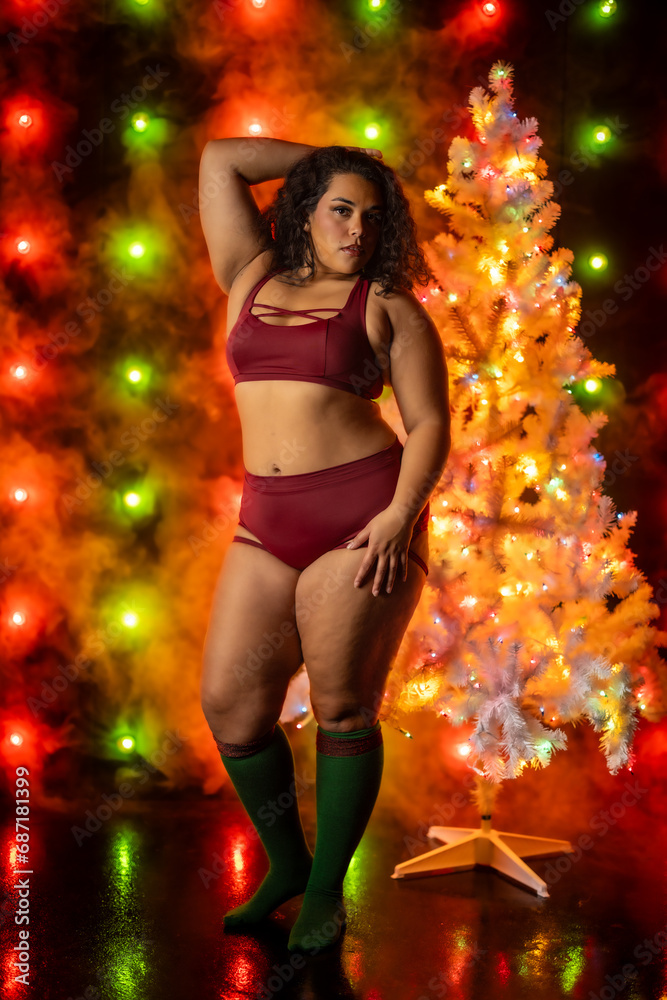 Fototapeta premium Female in sports bra and short posing in front of red and green christmas themed light wall next to white christmas tree