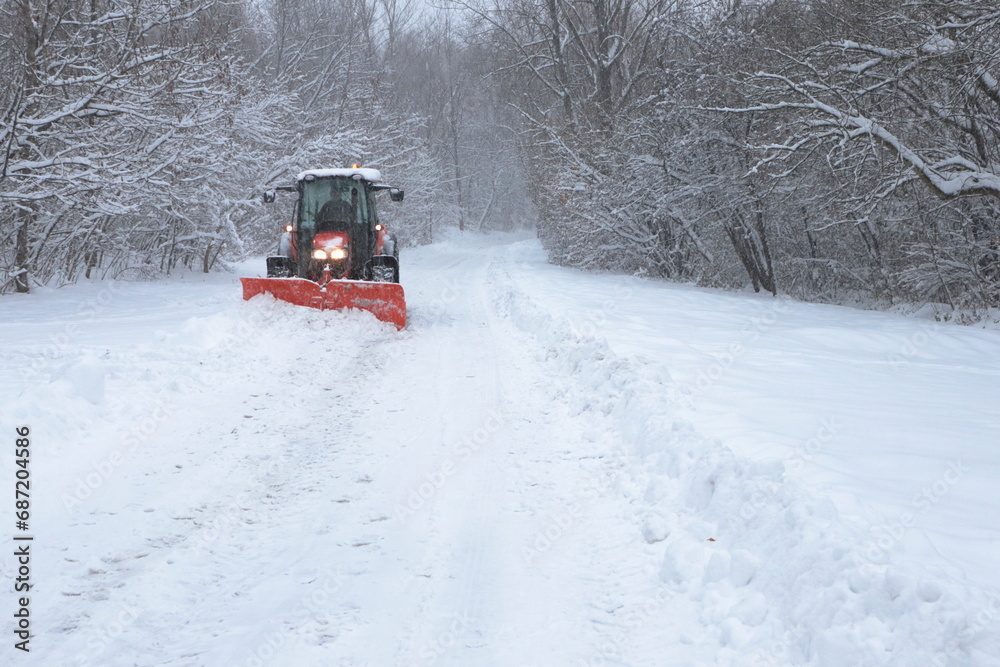 Tractor with snow plow clearing a forest road after heavy snowfall. Winter maintenance vehicle working in snowy landscape surrounded by trees Tractor with snow plow clearing a forest road after heavy snowfall. Winter maintenance vehicle working in snowy landscape surrounded by trees