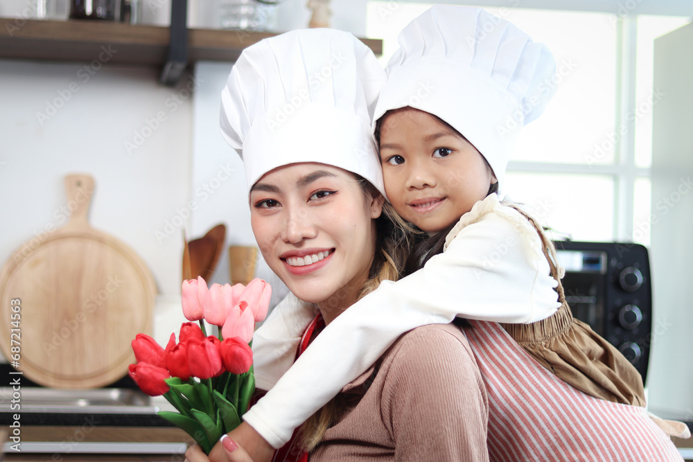 Smiling Asian mother and daughter wear apron chef hat at kitchen, girl giving tulip flowers and hugging her mom. Mother piggybacking kid, lovely family cooking food at home together. Happy mother day.