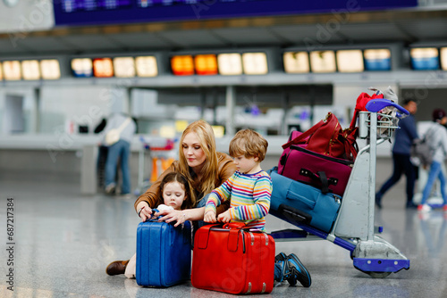 Two tired little kids, boy and girl, siblings and mother at the airport. Children, family traveling, going on vacation by plane and waiting by luggage trolley with suitcases at terminal for flight. photo