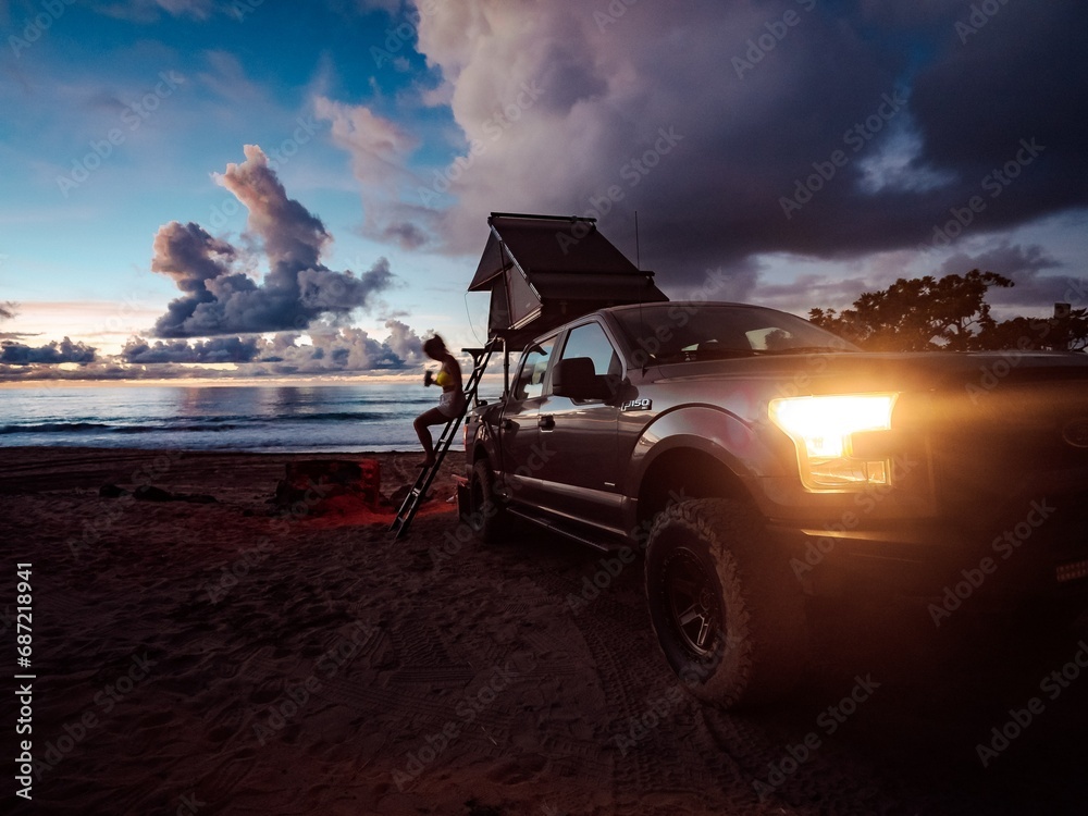 As the sun sets over the golden sand, a fearless woman perches on a ladder atop a truck, her jeep and its wheels ready to take her on a wild journey through the hawaiian islands of kauai and oahu, wi - obrazy, fototapety, plakaty 