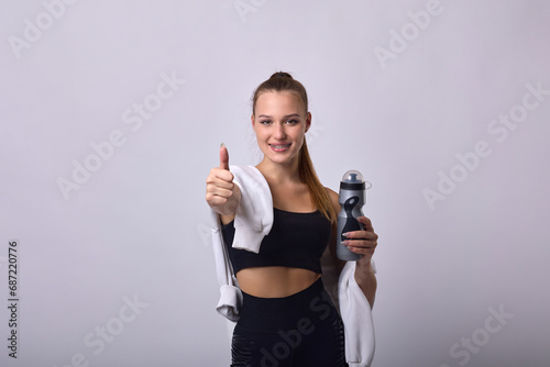 Sporty young woman drinking water from a bottle, standing on a gray background in a black suit. Portrait of sweaty slender girl taking a break after an intense workout in the gym © Aleksandr