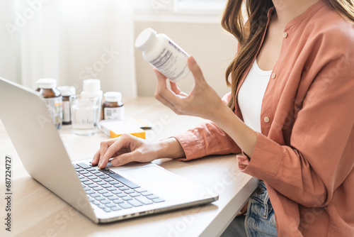 Wellness and dieting asian young woman, girl working from home using computer, typing or searching prescription on medicine label about vitamins information online, holding bottle of food supplement. photo
