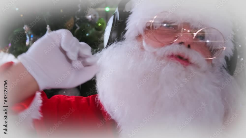 Close-up of gray-bearded Santa wearing disco dancer headphones waving his arms against backdrop of Christmas Tree decorated with toys and sparkling garlands. New Year party.
