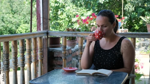Young caucasain woman drinking tea or coffe and reading book on summer terrace, slow motion photo