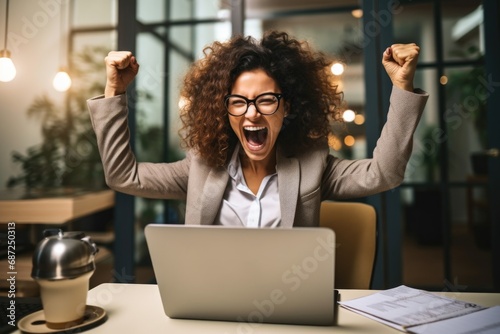 A professional woman in a suit and glasses raises her arms in the air in front of a laptop. 