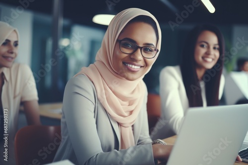 A woman wearing a hijab smiles as she works on her laptop. This image can be used to depict diversity in the workplace and the empowerment of women