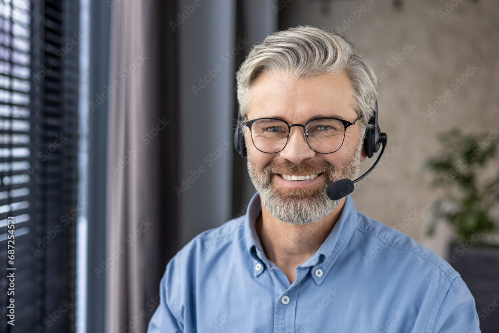 Close up portrait of mature senior man with headset phone for video call, businessman smiling and looking at camera, boss inside office at workplace. - obrazy, fototapety, plakaty 