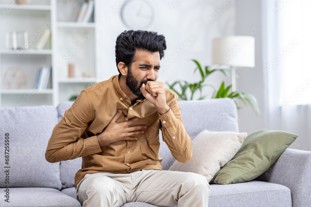 Indian young man sitting at home on the couch and coughing, covering his mouth, holding his chest, feeling severe pain, suffering from an infectious disease.