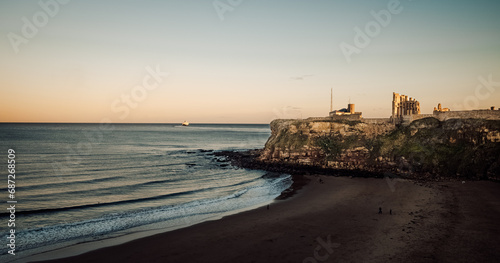 As the sun rises over the wild ocean, a lone boat floats towards the distant horizon, framed by the rugged cliff and sandy shore of the untamed beach