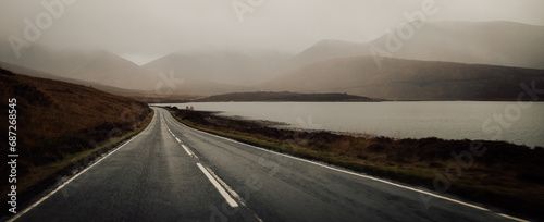 A winding highway cuts through the misty highlands, leading to a tranquil lake nestled between towering mountains and a vast open sky, inviting travelers to embark on a journey of discovery and awe i photo