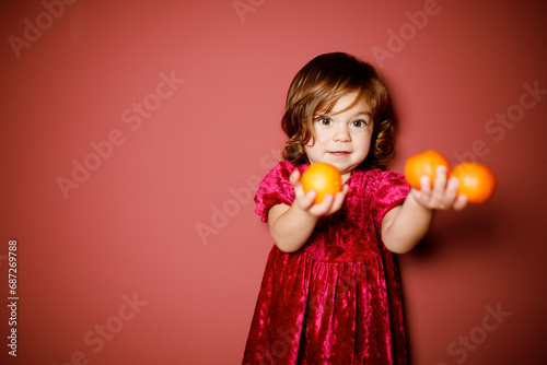 Adorable caucasian brunette toddler little girl in red velour  holding mandarin oranges in hands at the red background. Christmas vibe 