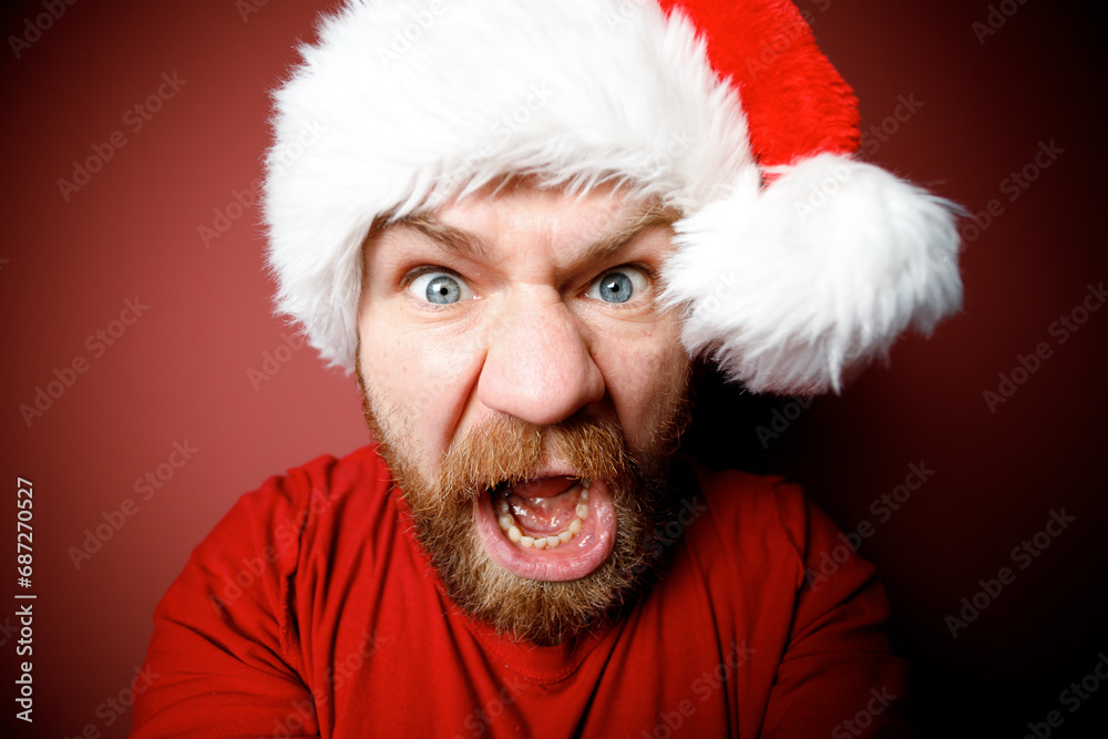 Portrait of grimacing young bearded Santa Claus in red hat with angry emotion with opened mouth, emotion faces on the red background Portrait of grimacing young bearded Santa Claus in red hat with angry emotion with opened mouth, emotion faces on the red background