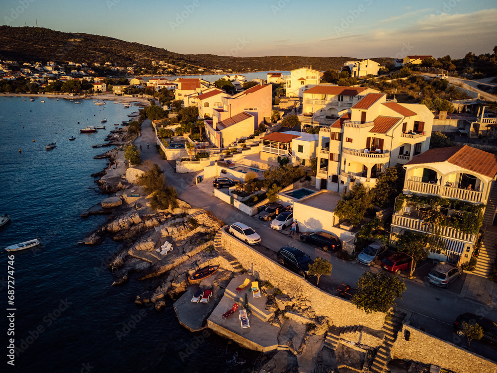 Fototapeta premium Amidst the tranquil landscape of a beachfront harbor, a row of colorful houses bask in the warm glow of a fiery sunset, as boats sway gently in the calm waters and the sky above transforms into a can