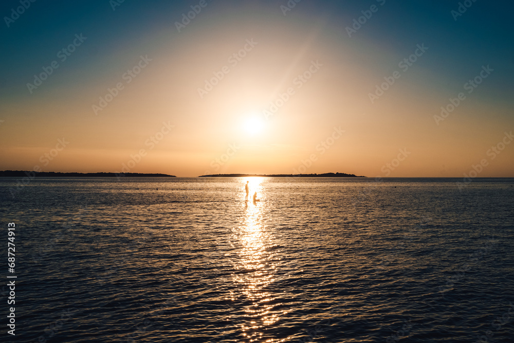 A lone figure drifts upon the tranquil waters, basking in the warm afterglow of a vibrant sunset, as the distant horizon melts into the golden sky and the soothing sound of gentle waves serenades the