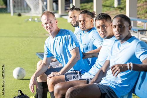 Group of 5 men on a soccer team wearing blue jerseys. The players are sitting together on a bench on the side of the game field. 