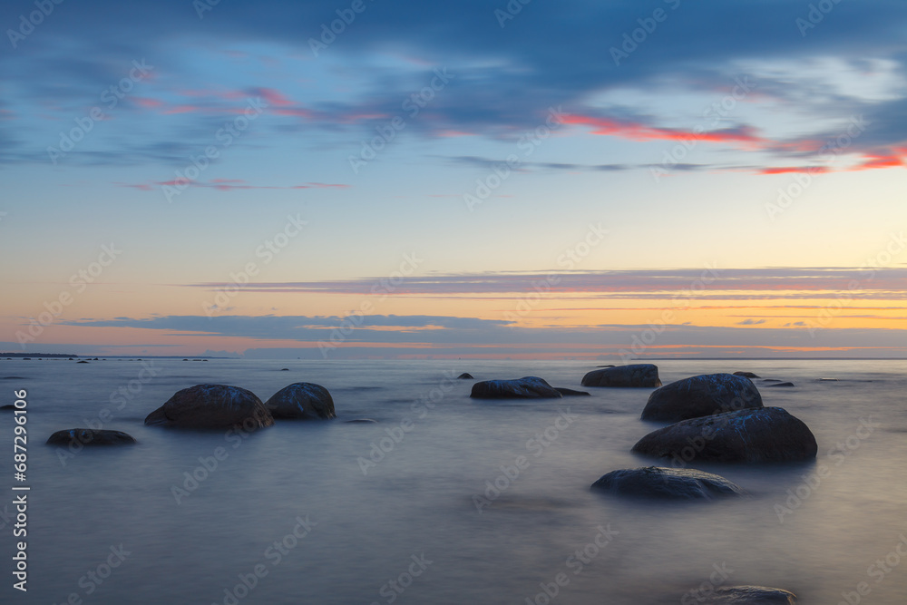 Romantic atmosphere in peaceful sunset at sea. Big boulders sticking out from smooth wavy sea. Yellow strip on horizon and tiny clouds