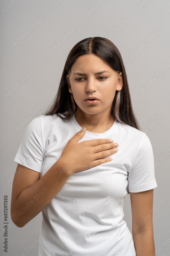 Teen girl feeling relief touching chest by hand looking down with calm face. Upset teenager suffer from breathing problems, panic attack, suffocation holding hand on chest isolated on gray background Teen girl feeling relief touching chest by hand looking down with calm face. Upset teenager suffer from breathing problems, panic attack, suffocation holding hand on chest isolated on gray background