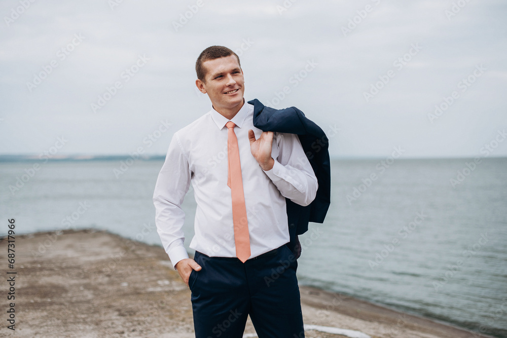 Handsome groom in a black suit with a red tie near sea. Wedding portrait of newlyweds. Handsome groom in a black suit with a red tie near sea. Wedding portrait of newlyweds.