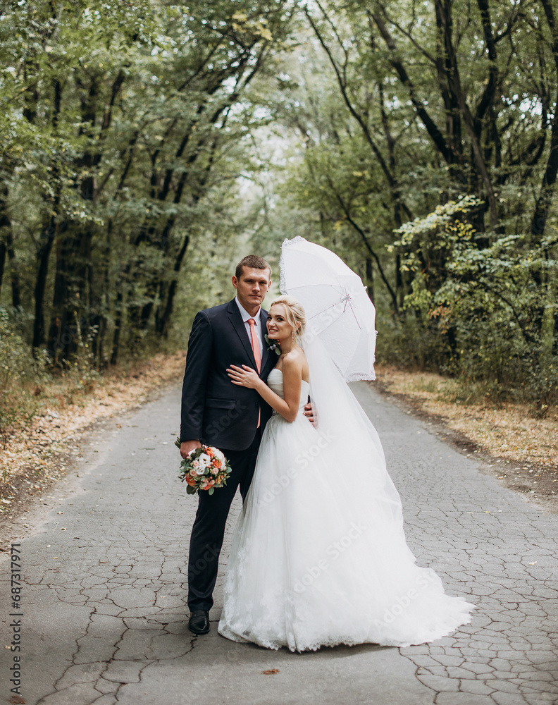 Beautiful wedding couple embracing together under umbrella in forest. Stylish bride and groom kissing