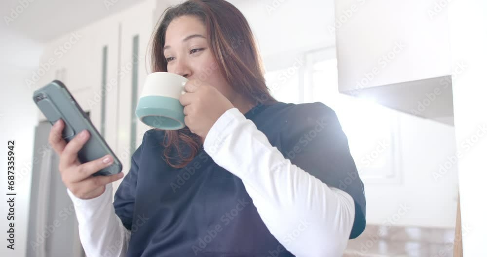 Happy plus size biracial female doctor having coffee using smartphone at home, slow motion Happy plus size biracial female doctor having coffee using smartphone at home, slow motion