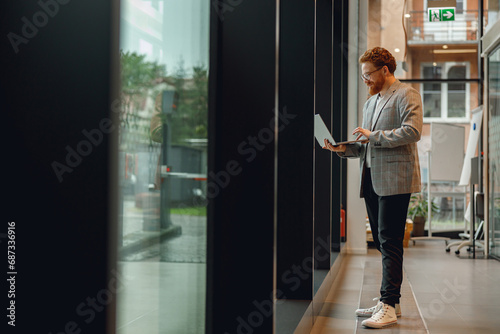 Focused male sales manager in eyeglasses working on laptop standing in office during working day