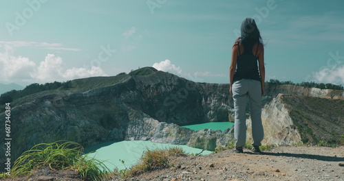 Woman tourist walking to Kelimutu blue crater lakes mountain edge. Back view of long hair girl in trekking clothes with hair blowing on wind. Travel, tourism, holiday, hiking, relax. photo