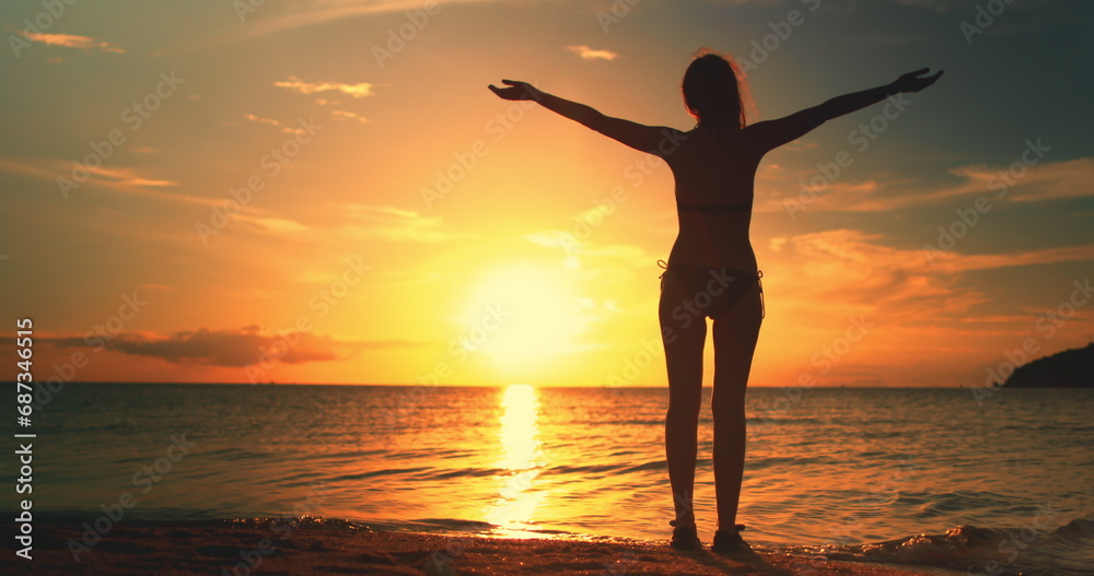 Woman enjoys amazing colorful sunset, standing on a tropical beach. Slim girl silhouette in bikini raises hands against bright orange dramatic sky and ocean waves. Holiday, travel, recreation concept Woman enjoys amazing colorful sunset, standing on a tropical beach. Slim girl silhouette in bikini raises hands against bright orange dramatic sky and ocean waves. Holiday, travel, recreation concept